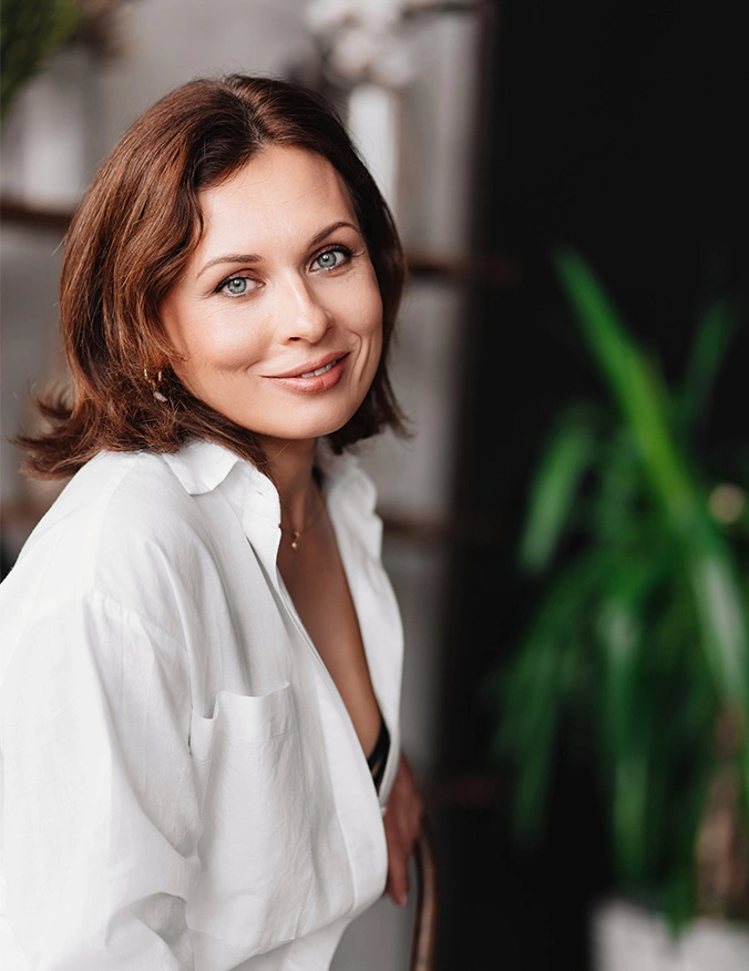 Woman with brown hair in white shirt smiling at camera in bright indoor setting with green plants - Bioidentical hormone replacement therapy in Newport Beach, CA
