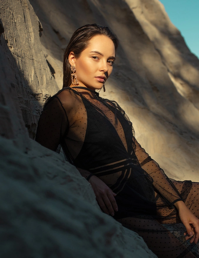 Woman in elegant black sheer outfit posing against rocky coastal landscape at golden hour