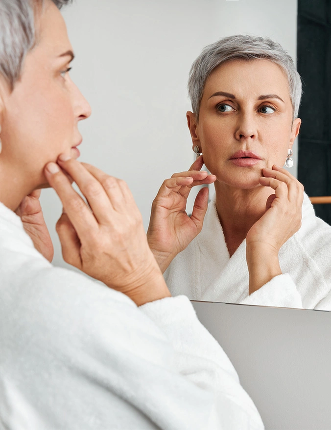 Woman with short gray hair examining her face in bathroom mirror while touching her chin - earlobe lift in Newport Beach, CA