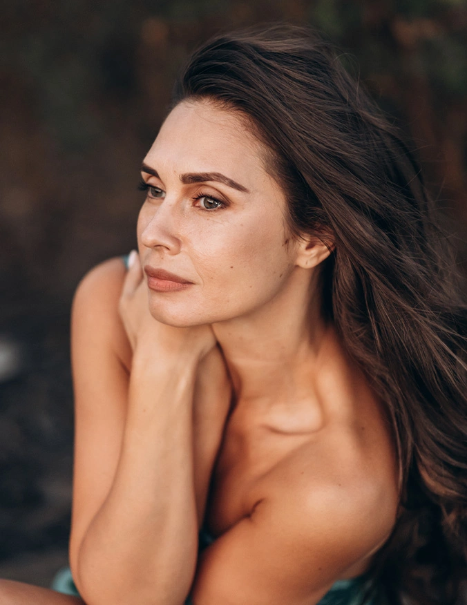 Beautiful woman with long dark hair posing elegantly with hands near face in natural outdoor lighting - fat transfer into face in Newport Beach, CA