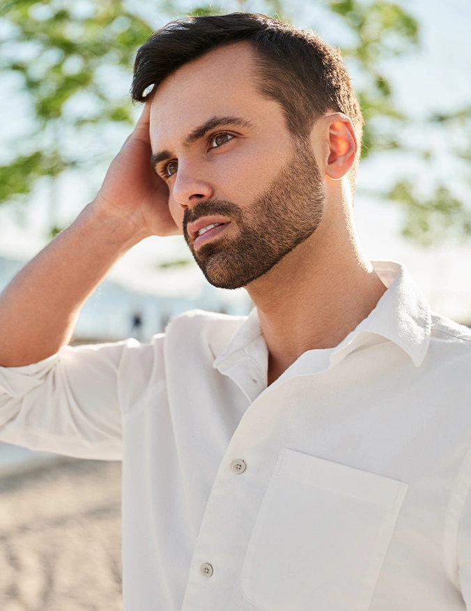 Handsome bearded man in white shirt touching hair outdoors by beach on sunny day - otoplasty in Newport Beach, CA