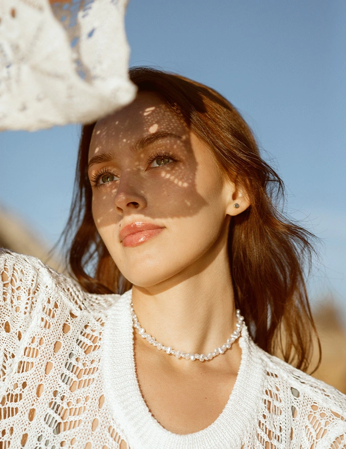 Woman in white lace top and pearl necklace enjoying sunny day outdoors with natural lighting and blue sky - Rhinoplasty in Newport Beach, CA