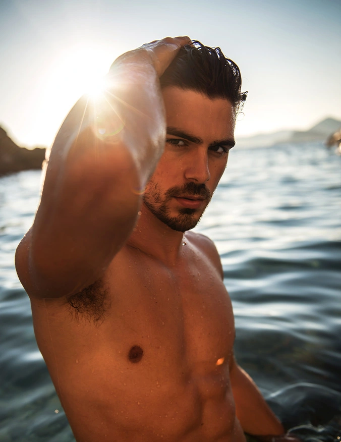 Shirtless man with hand in hair by water at sunset with sun flare and ocean backdrop - Gynecomastia in Newport Beach, CA