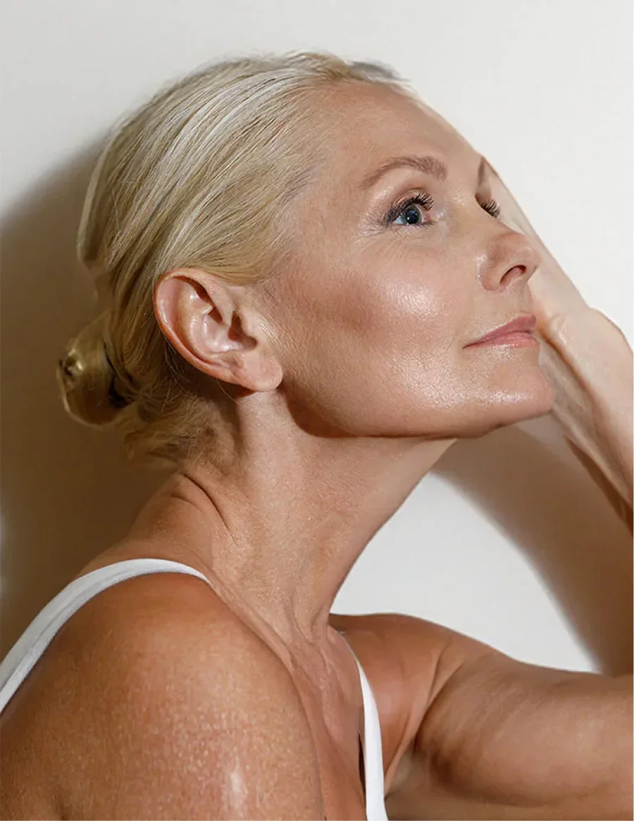 Mature woman with natural aging skin touching face in white tank top against neutral background