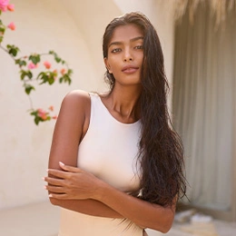 Woman with long dark hair in white tank top posing elegantly against beige curtain backdrop with flowers - Brow lift in Newport Beach, CA