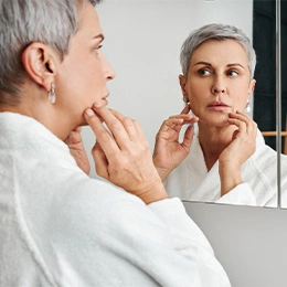 Mature woman examining facial skin in mirror while touching face in white bathrobe - Earlobe lift in Newport Beach, CA