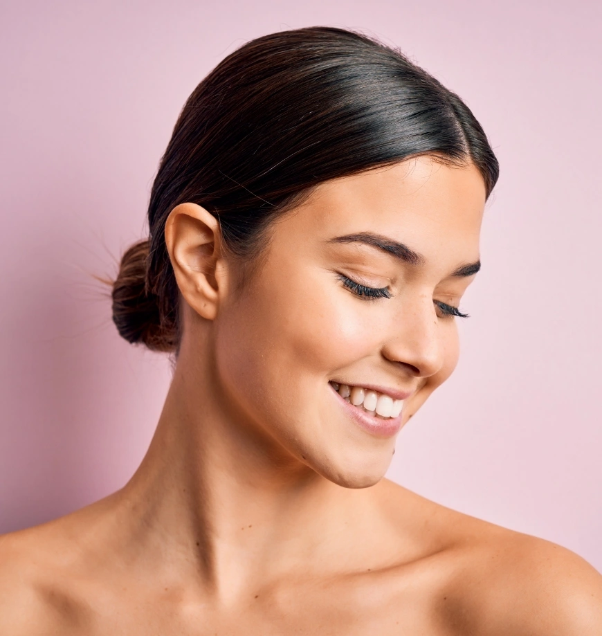 Woman with smooth, glowing skin and elegant updo hairstyle smiling against pink background - facials in Newport Beach, CA