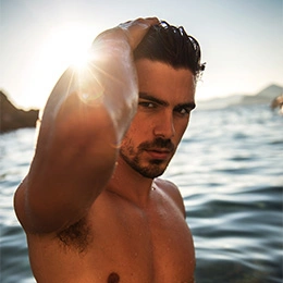 Shirtless man posing by water at sunset with hand in hair, beach lifestyle portrait - fat transfer breast augmentation in Newport Beach, CA