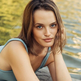 Young woman with brown hair in blue tank top sitting outdoors by water on sunny day - Lip augmentation in Newport Beach, CA
