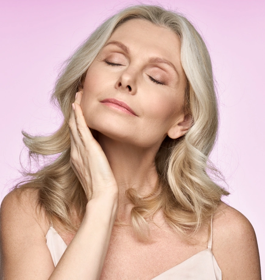 Mature woman with blonde hair touching face showing healthy glowing skin against pink background - morpheus8 in Newport Beach, CA